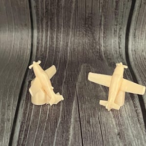 May include: Two small, light-colored airplane models are displayed on a wooden surface. One plane appears to be damaged, while the other is intact. The planes have wings and tails, and are likely toys or decorative items.
