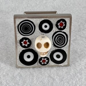 May include: A silver-toned rectangular box with a skull and black and white circular and floral designs. The skull is centered, with a white base and brown eye sockets. The box has a silver hinge.