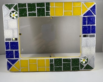 May include: A rectangular mosaic picture frame with a clear glass insert. The frame features a colorful design with green, yellow, blue, and silver tiles. Small white flower accents are placed on the corners. The frame is designed to hold a photograph.