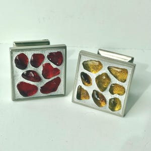 May include: Two square cufflinks with a silver metal frame. One has red, irregularly shaped stones, the other amber-coloured stones. The cufflinks are set against a light background. Each cufflink has a metal bar.