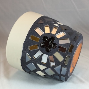 Puede incluir: Un macetero de cerámica gris con un diseño de mosaico que presenta azulejos espejados y una flor negra en el centro. El macetero tiene una base blanca y un borde marrón.