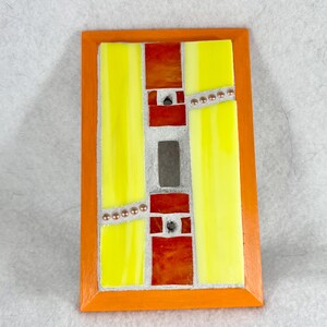 May include: A rectangular stained glass art piece with an orange wooden frame. The design features vertical yellow and red glass panels, accented with small, round, copper-colored beads. The central section is a silver-colored rectangle.
