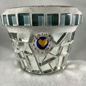 May include: A handcrafted mosaic flower pot with a textured, light gray exterior. The rim features teal square tiles, while the body is adorned with mirrored glass pieces. A blue and gold heart-shaped accent adds a decorative touch.