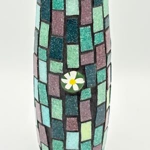 May include: A mosaic vase with a cylindrical shape. The vase is covered in small, rectangular tiles in shades of green, teal, and purple. A small, round, white and yellow flower is affixed to the front of the vase. The vase has a dark grout between the tiles.