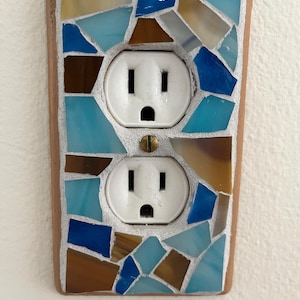 May include: A white electrical outlet with two plugs is surrounded by a mosaic tile cover. The tiles are in shades of blue, brown, and beige.