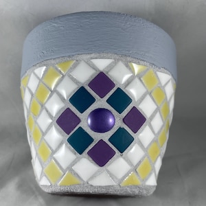 Puede incluir: Un pequeño macetero de cerámica gris con un diseño de mosaico. El mosaico está hecho de azulejos blancos, amarillos, morados y azul verdoso. El macetero tiene un solo azulejo morado en el centro.