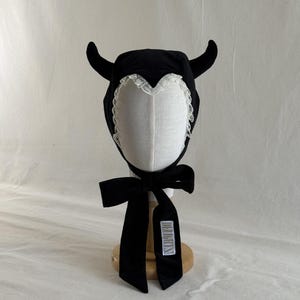 Puede incluir: Un gorro negro con dos cuernos curvos en los lados, un borde de encaje blanco a lo largo de la abertura facial y una cinta negra larga para atar bajo la barbilla. Tiene una etiqueta rectangular blanca con texto negro en la cinta.