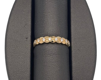 May include: A gold ring with a repeating pattern of diamond-shaped cutouts, each containing a small, clear gemstone. The ring is displayed on a black jewelry stand against a white background. The ring's design is symmetrical and elegant.
