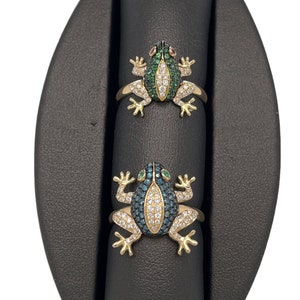 May include: Two gold-toned frog rings. The frogs have green, white, and blue gemstone details. The rings are displayed on a black jewelry stand. The frog's eyes are red and green gemstones.