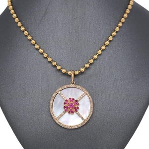 Pode incluir: Colar dourado com pingente redondo. O pingente apresenta um centro de madrepérola, um grupo de pedras preciosas rosa e uma borda cravejada de diamantes. O colar tem uma corrente de contas.