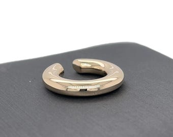 Könnte beinhalten: Ein goldfarbener Ohrklemme mit einem glatten, abgerundeten Design. Der Schmuck hat eine C-Form und ist an einem Ende offen. Der Schmuck wird auf einer dunkelgrauen Oberfläche präsentiert, was einen Kontrast erzeugt, der den metallischen Glanz des Ohrschmucks hervorhebt.