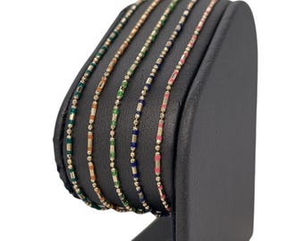 May include: A set of seven gold bracelets with colourful enamel accents. The bracelets are displayed on a black stand.
