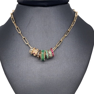 Könnte beinhalten: Eine goldene Kettenkette mit einem Anhänger aus mehreren Ringen, die mit grünen, roten und blauen Edelsteinen besetzt sind.