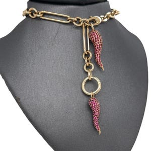 May include: Gold-coloured necklace with a chain and a pendant featuring two red horn charms. The charms are encrusted with small, sparkling stones. The necklace is displayed on a black mannequin.