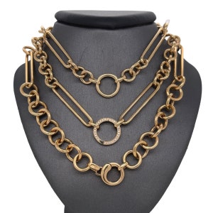 May include: A gold layered necklace with three different chain styles. The top chain has a large circle pendant, the middle chain has a smaller circle pendant with small diamonds, and the bottom chain has a large circle clasp.