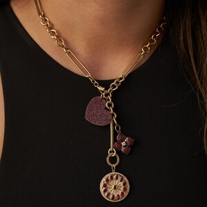 May include: A gold chain necklace with a heart, flower, and round charm pendant. The charms are encrusted with red gemstones.