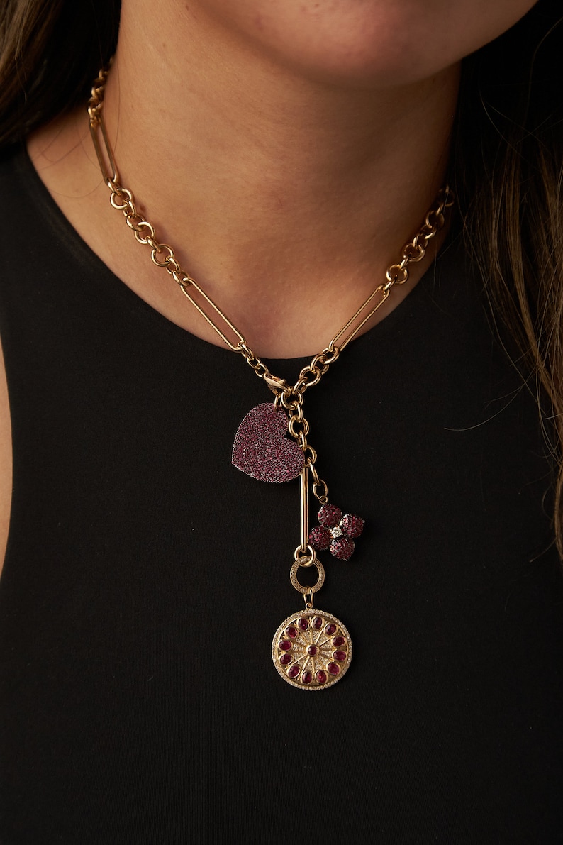 May include: A gold chain necklace with a heart, flower, and round charm pendant. The charms are encrusted with red gemstones.