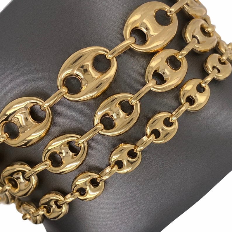 K&ouml;nnte beinhalten: Nahaufnahme von drei goldfarbenen Kettenarmb&auml;ndern. Jedes Armband hat eine Reihe von ovalen Gliedern, die durch kleinere Glieder verbunden sind. Die Armb&auml;nder werden auf einer grauen Oberfl&auml;che pr&auml;sentiert, die ihre gl&auml;nzende, reflektierende Oberfl&auml;che zeigt.