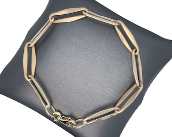 Peut inclure: Un bracelet à maillons de couleur or avec un design ovale. Le bracelet est présenté sur une surface gris foncé. Le fermoir est visible et les maillons sont reliés en chaîne.