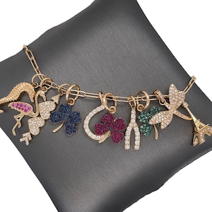 Könnte beinhalten: Ein goldfarbenes Charm-Armband mit verschiedenen Anhängern, darunter ein Flamingo, ein Hufeisen, ein vierblättriges Kleeblatt, ein Seepferdchen, ein Wunschknochen, eine Libelle und der Eiffelturm. Jeder Anhänger ist mit funkelnden Edelsteinen verziert.