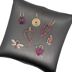 Puede incluir: Una colección de colgantes dorados con varios diseños, incluyendo una mariquita, un ojo, un corazón, una libélula, un candado y un dije de "I ❤️ NY". Los colgantes están adornados con piedras preciosas rosas y transparentes.