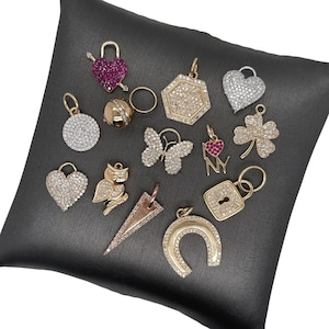 May include: A collection of gold-toned and silver-toned charms, including hearts, a lock, a horseshoe, a butterfly, a four-leaf clover, and an owl. Some charms are encrusted with small, sparkling stones. The charms are displayed on a dark gray surface.