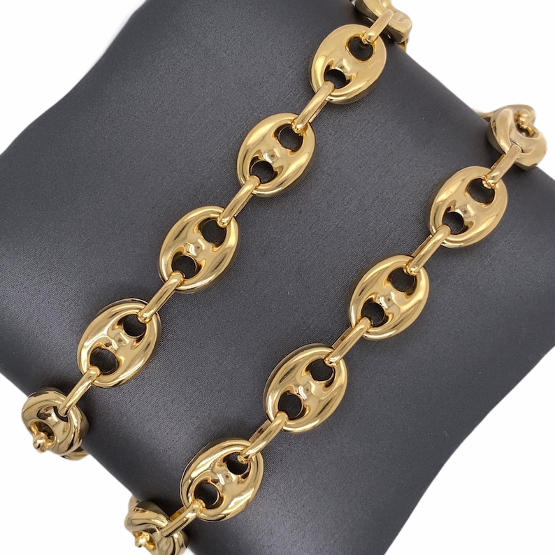 K&ouml;nnte beinhalten: Zwei goldfarbene Kettenarmb&auml;nder mit einem markanten, aufgebl&auml;hten ovalen Gliederdesign. Die Glieder sind durch kleinere, l&auml;ngliche Glieder verbunden. Die Armb&auml;nder werden auf einer dunkelgrauen Oberfl&auml;che pr&auml;sentiert, die ihre gl&auml;nzende, reflektierende Oberfl&auml;che hervorhebt.