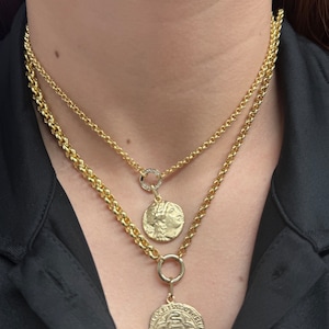 May include: Two gold chain necklaces with a coin pendant on each. The top chain has a small circle pendant with a single diamond. The bottom chain has a larger coin pendant with an intricate design.