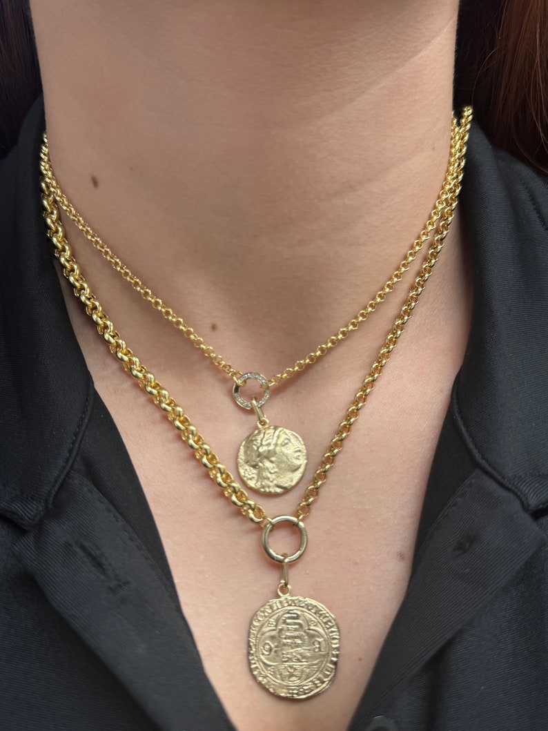 May include: Two gold chain necklaces with a coin pendant on each. The top chain has a small circle pendant with a single diamond. The bottom chain has a larger coin pendant with an intricate design.