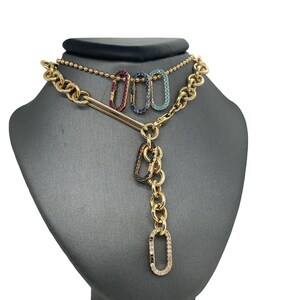 May include: Gold-toned necklace with a chain and a pendant featuring three colored, gem-studded oval charms: red, blue, and turquoise. The pendant has a vertical chain of gold links with a diamond-studded oval at the bottom.