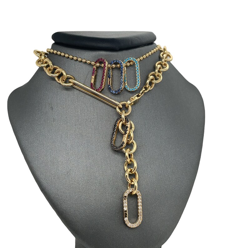 May include: Gold-toned necklace with a chain and a pendant featuring three colored, gem-studded oval charms: red, blue, and turquoise. The pendant has a vertical chain of gold links with a diamond-studded oval at the bottom.
