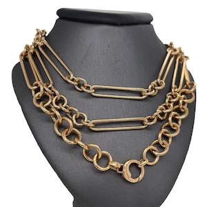 May include: A gold-toned layered chain necklace with a lobster clasp closure. The necklace features three different chain styles, including a thick curb chain, a thin curb chain, and a chain with rectangular links.