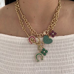 May include: A gold chain necklace with multiple charms. The charms are a green heart, a green four-leaf clover, a red horseshoe, and a red flower. The charms are all covered in sparkling stones.