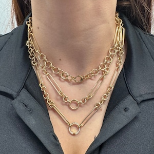 May include: A layered gold chain necklace with a small, round pendant set with clear stones. The necklace features three different chain styles, including a chunky curb chain, a delicate chain, and a chain with rectangular links.