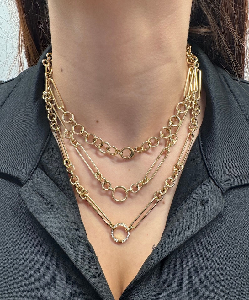 May include: A layered gold chain necklace with a small, round pendant set with clear stones. The necklace features three different chain styles, including a chunky curb chain, a delicate chain, and a chain with rectangular links.