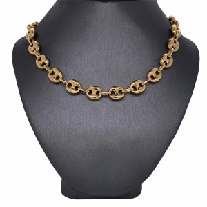 K&ouml;nnte beinhalten: Eine goldfarbene Halskette mit einem Kaffeebohnen-Kettendesign. Die Halskette wird auf einer schwarzen Schmuckb&uuml;ste pr&auml;sentiert. Die Kettenglieder sind oval geformt und haben eine zentrale Vertiefung, was ein stilvolles Accessoire ergibt.