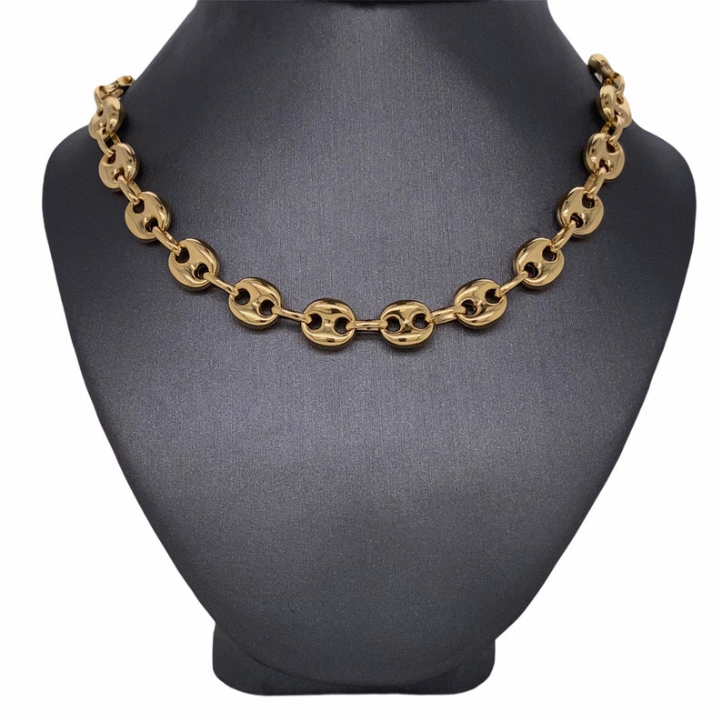 K&ouml;nnte beinhalten: Eine goldfarbene Halskette mit einem Kaffeebohnen-Kettendesign. Die Halskette wird auf einer schwarzen Schmuckb&uuml;ste pr&auml;sentiert. Die Kettenglieder sind oval geformt und haben eine zentrale Vertiefung, was ein stilvolles Accessoire ergibt.