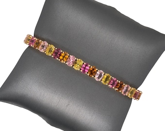 May include: A gold bracelet with a row of colorful oval-shaped gemstones. The gemstones are pink, orange, yellow, and green.