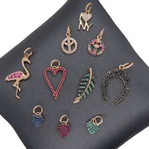 May include: A collection of 10 gold charm pendants with colorful gemstones. The charms include a peace sign, a heart, a horseshoe, a leaf, a flamingo, and the letters "NY".