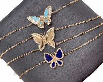 Könnte beinhalten: Drei goldene Schmetterlingsketten. Ein Schmetterling hat ein hellblaues und weißes Design, ein anderer ist goldfarben und der dritte ist dunkelblau. Jeder Schmetterling ist mit kleinen, funkelnden Steinen verziert.