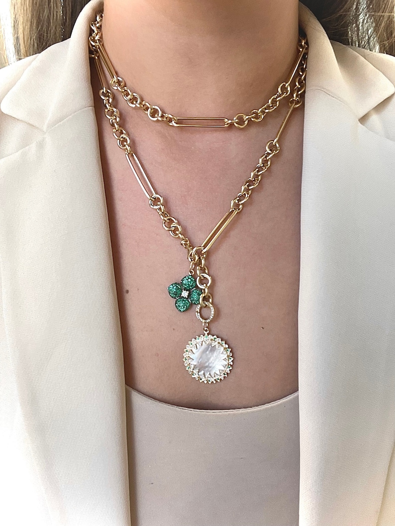 May include: A gold chain necklace with a layered design. The necklace features a pendant with a green flower charm and a round, white, mother-of-pearl charm.