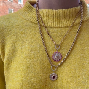 May include: A gold chain necklace with two pendants. The top pendant is a round circle with a smaller circle in the center. The bottom pendant is a round disc with a face in the center. The face is surrounded by pink gemstones.