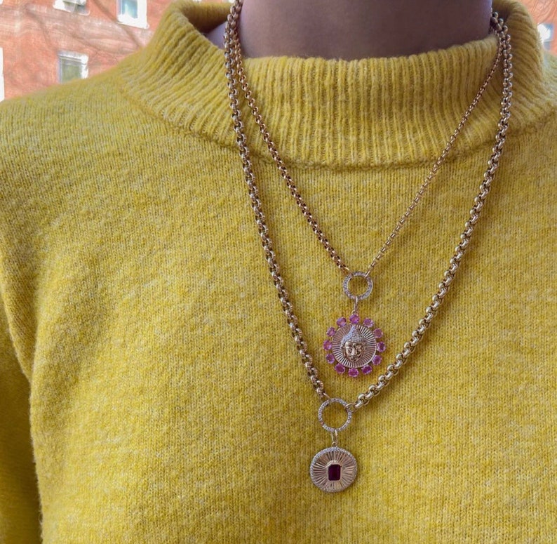 May include: A gold chain necklace with two pendants. The top pendant is a round circle with a smaller circle in the center. The bottom pendant is a round disc with a face in the center. The face is surrounded by pink gemstones.