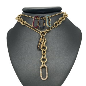 Puede incluir: Collar dorado con una variedad de estilos de colgantes. El collar presenta una cadena con una barra y un cierre, y una cadena colgante de anillos. Los colgantes incluyen tres pequeños cierres de mosquetón de colores con gemas y dos cierres ovalados más grandes.