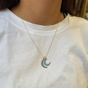 K&ouml;nnte beinhalten: Eine goldene Kettenkette mit einem Anh&auml;nger in Form eines doppelten Halbmondes. Der Mond ist mit Gold umrandet und mit hellblauem Email gef&uuml;llt.