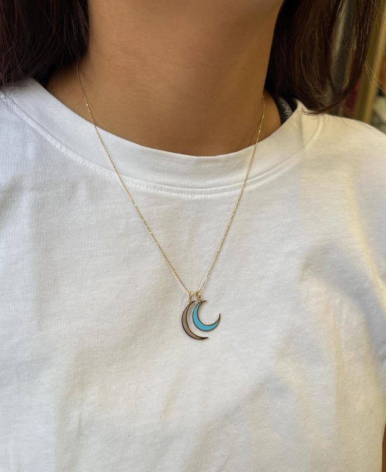 K&ouml;nnte beinhalten: Eine goldene Kettenkette mit einem Anh&auml;nger in Form eines doppelten Halbmondes. Der Mond ist mit Gold umrandet und mit hellblauem Email gef&uuml;llt.