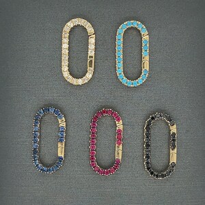 May include: Five gold-toned oval-shaped jewelry clasps. Each clasp is encrusted with gemstones in various colors: clear, turquoise, dark blue, red, and black. The clasps are displayed on a dark gray surface.