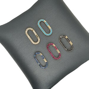 May include: Five oval-shaped jewelry clasps with various gemstone colors: clear, turquoise, black, ruby, and sapphire. Each clasp is set in a gold-tone metal and rests on a gray satin surface. The clasps are designed for jewelry making.