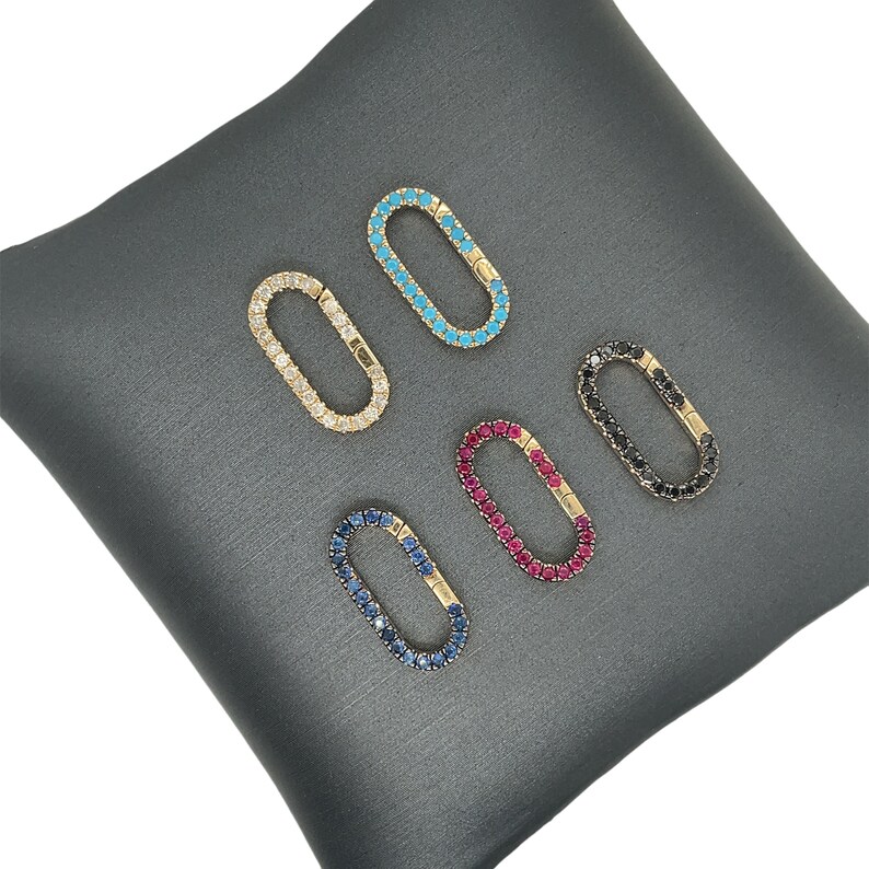 May include: Five oval-shaped jewelry clasps with various gemstone colors: clear, turquoise, black, ruby, and sapphire. Each clasp is set in a gold-tone metal and rests on a gray satin surface. The clasps are designed for jewelry making.