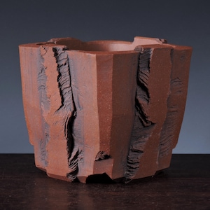 May include: A terracotta-colored ceramic pot with a wide, open top and a textured exterior. The surface features vertical, jagged, and deeply grooved panels that alternate with smooth, flat sections, creating a rugged, layered appearance.